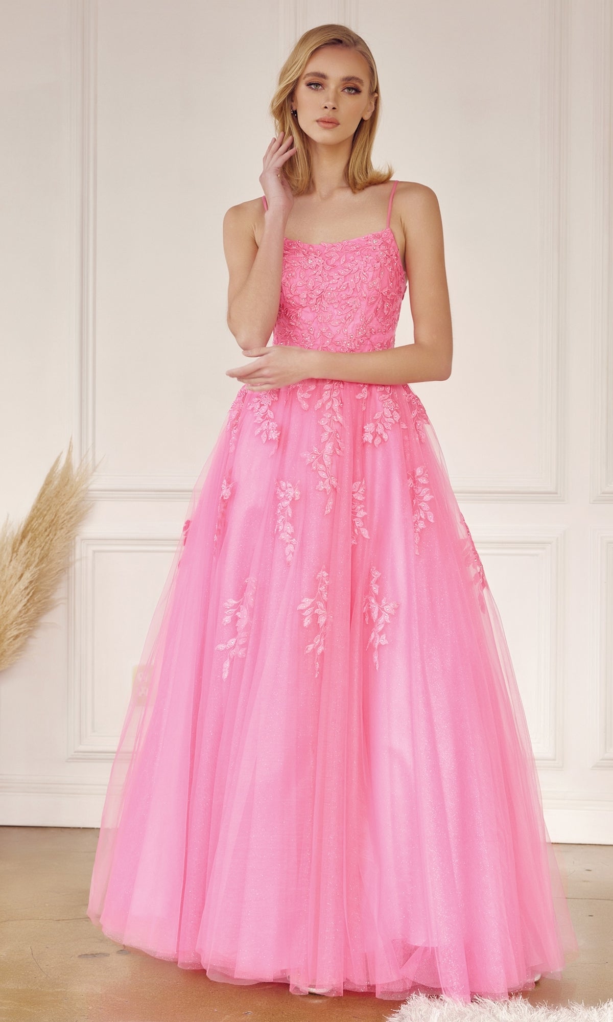 A woman poses indoors in the Corset-Back Long Glitter-Tulle Prom Ball Gown 260, featuring a long bright pink design with floral embroidery and thin straps, touching her face with one hand.